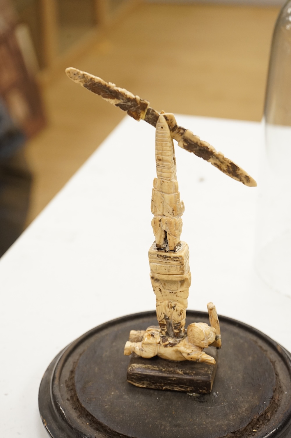 A 19th century Indian bone sculptural devotional object, carved as a totem with Ganesh and other deities, pivoting beam, 18cm high, glass dome with ebonised base